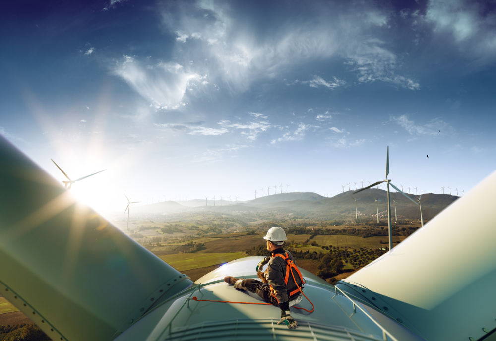 A worker in safety gear sitting on top of a wind turbine with a scenic view of multiple wind turbines in a green landscape under a partly cloudy sky.